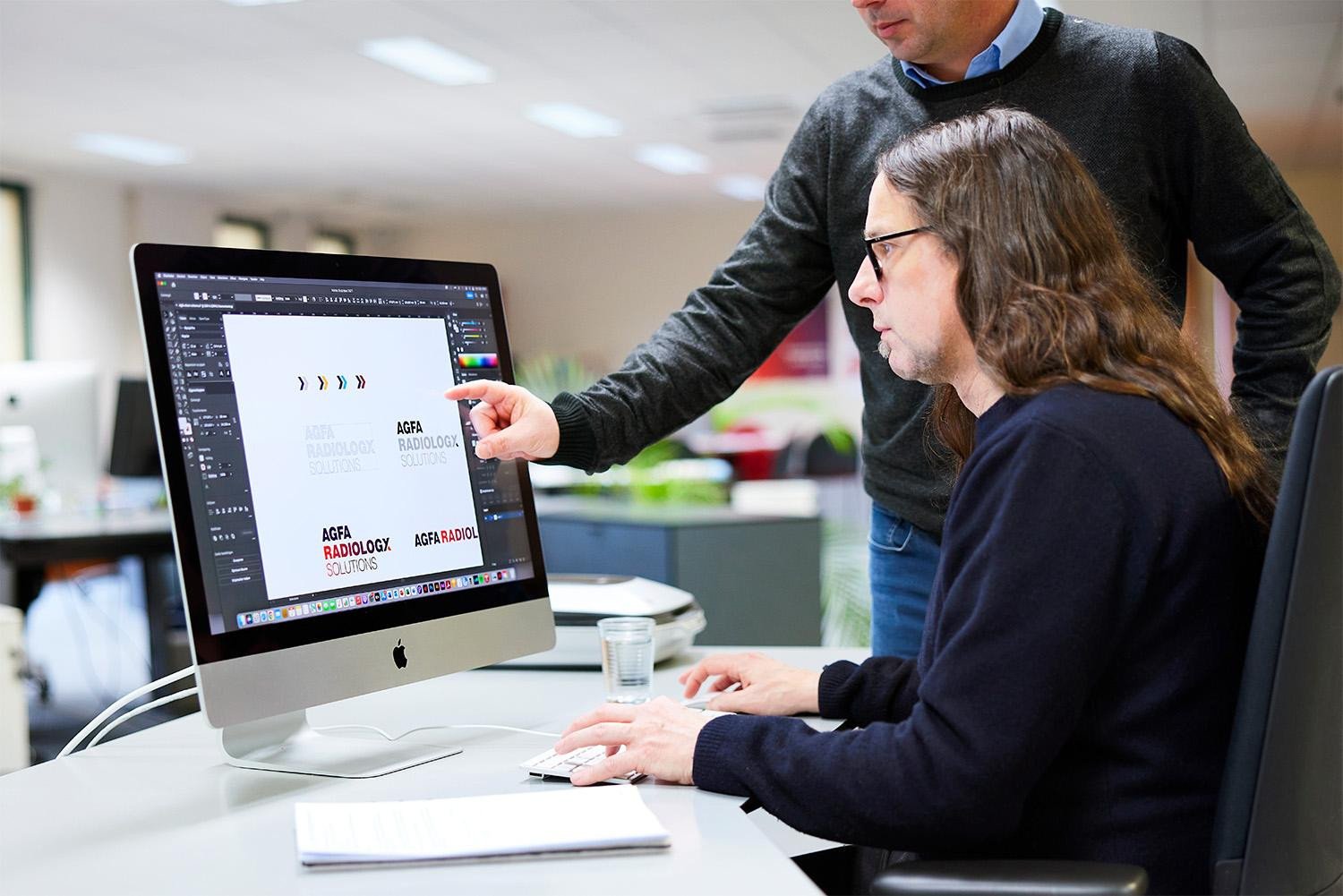 Designers bespreken logo-ontwerpen voor Agfa Radiology Solutions aan een iMac op kantoor.