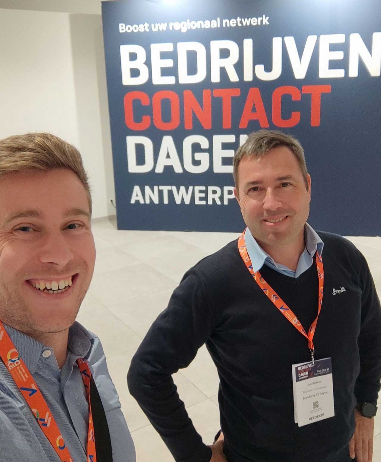 Twee mannen poseren tijdens de Bedrijvencontactdagen in Antwerpen, met een groot evenementendoek op de achtergrond.
