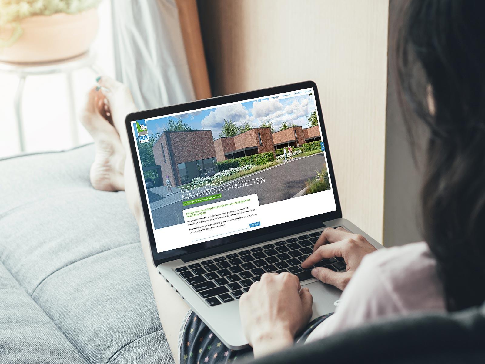 Laptop op schoot met website van RDK, waarop een nieuwbouwproject met moderne woningen en groene omgeving wordt gepresenteerd.