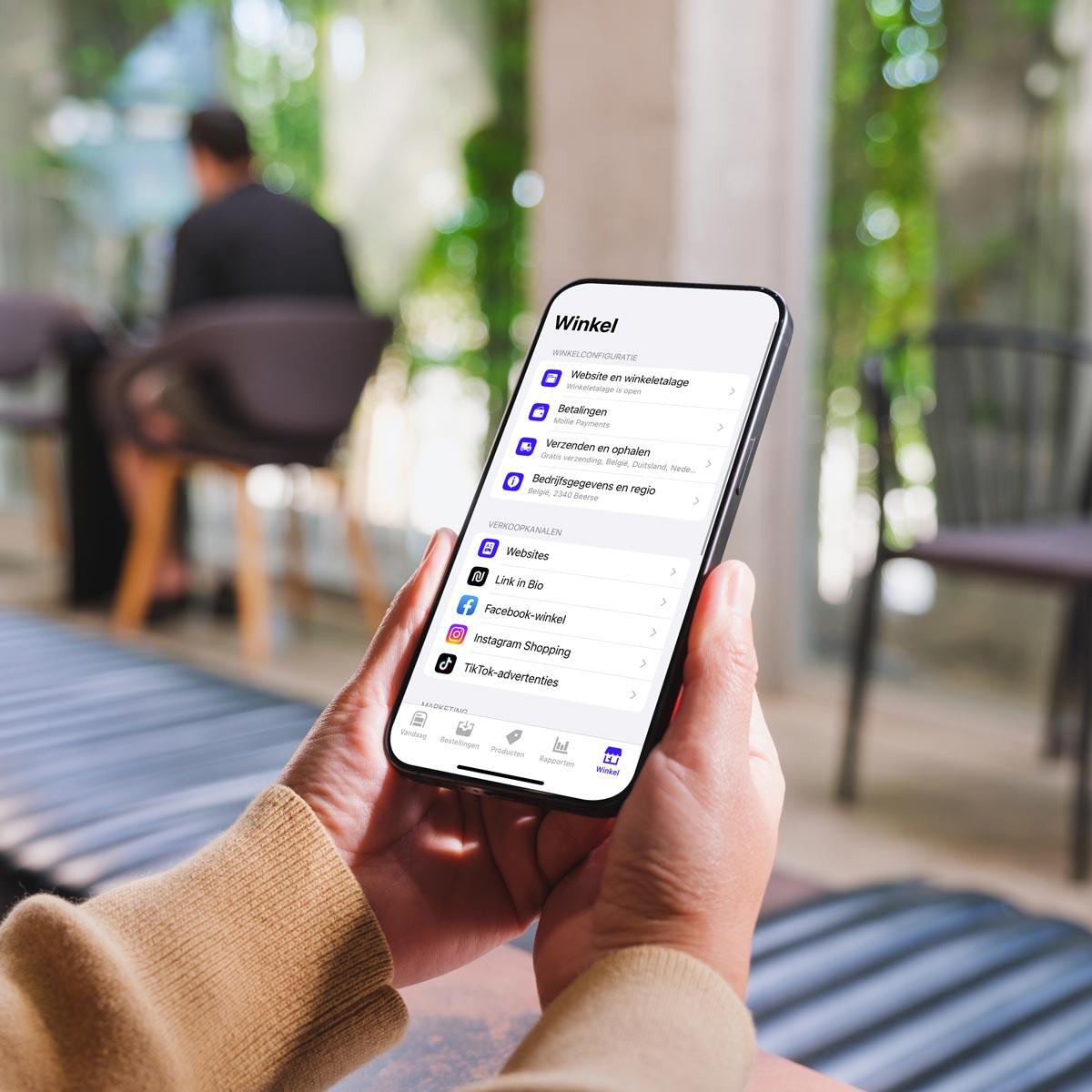 Handen houden een smartphone vast met de Ecwid winkel-app, waarop instellingen voor webshop, betalingen en verkoopkanalen zichtbaar zijn.